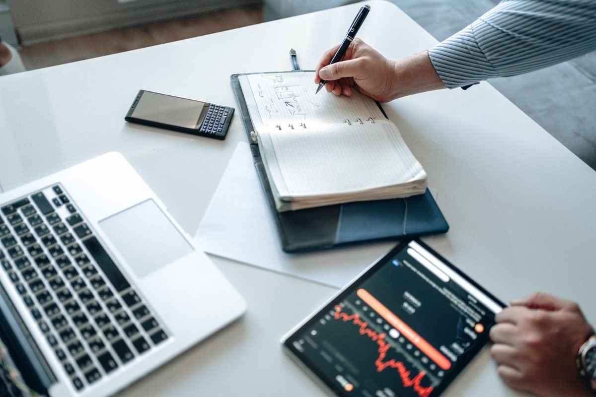 Anotando un plan de inversión en un cuaderno junto a un portátil con gráficos de bolsa